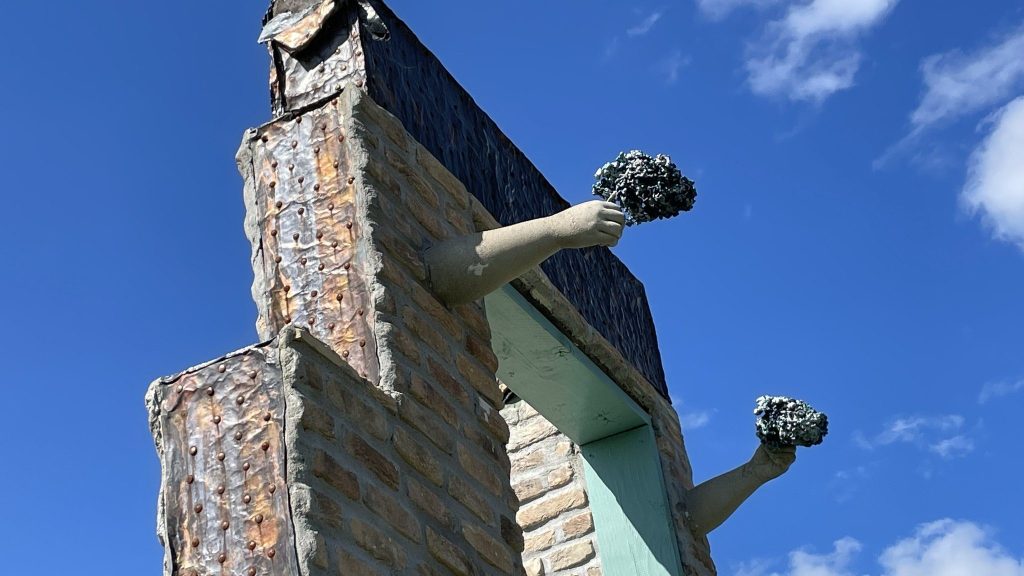 Stone sculpture of two figures with raised arms holding out flowers bright blue sky, symbolizing beauty in perseverance and achievement